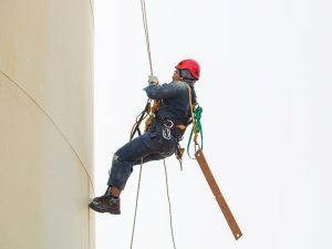 Nova NR35. Foto mostra um homem escalando um silo de cor branca. Ele usa equipamento de segurança e capacete vermelho