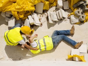 Investigação de Acidentes de trabalho. Foto mostra homem caído no chão e outro funcionário da empresa tentando ajudá-lo. Ambos estão com roupas amarelas e com capacete.